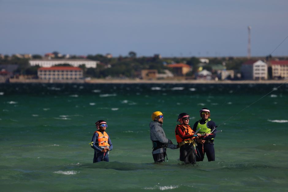 Four kite surfers in wetsuits and helmets training in the ocean near a waterfront.