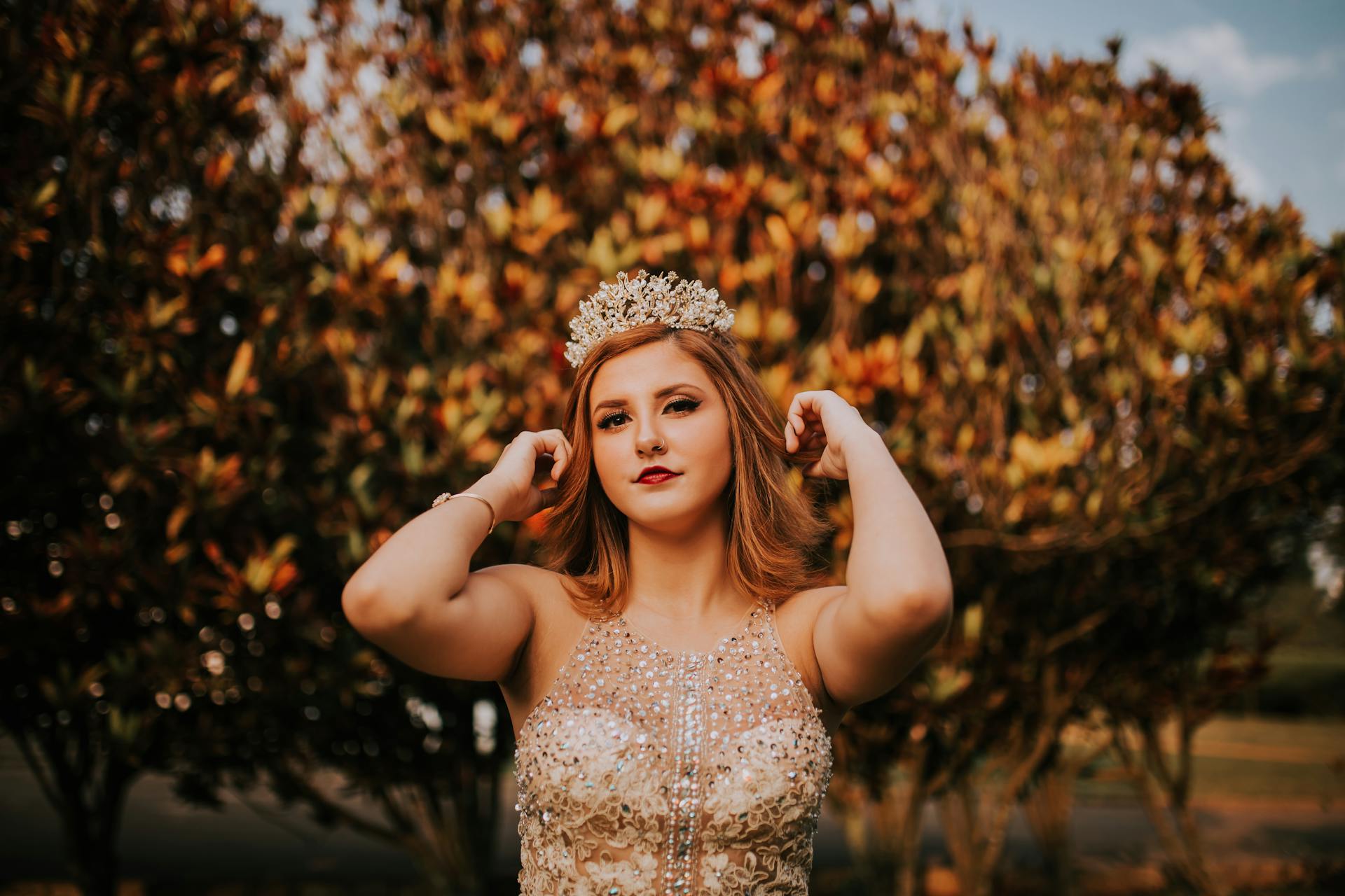 Beautiful woman in jeweled dress adjusts crown under autumn trees. Elegant and regal vibe.