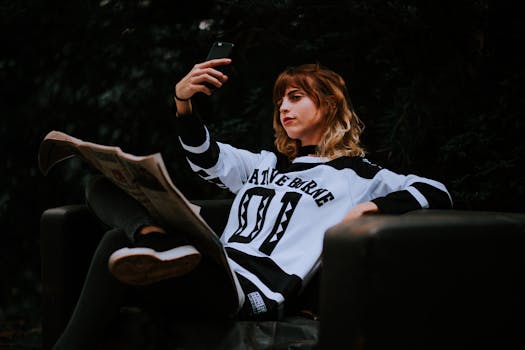 A teenage girl in a sports jersey taking a selfie outdoors while sitting on a bench during dusk.