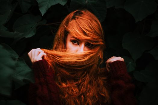 A captivating portrait of a woman with auburn hair partially covering her face, surrounded by lush green leaves.