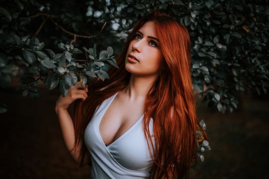A beautiful red-haired young woman posing gracefully outdoors among lush green leaves.