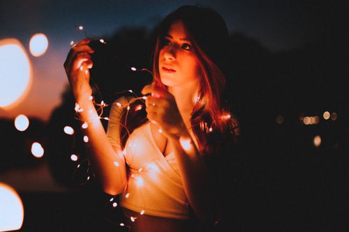 Free A young woman illuminated by string lights against a dark, moody evening backdrop. Stock Photo