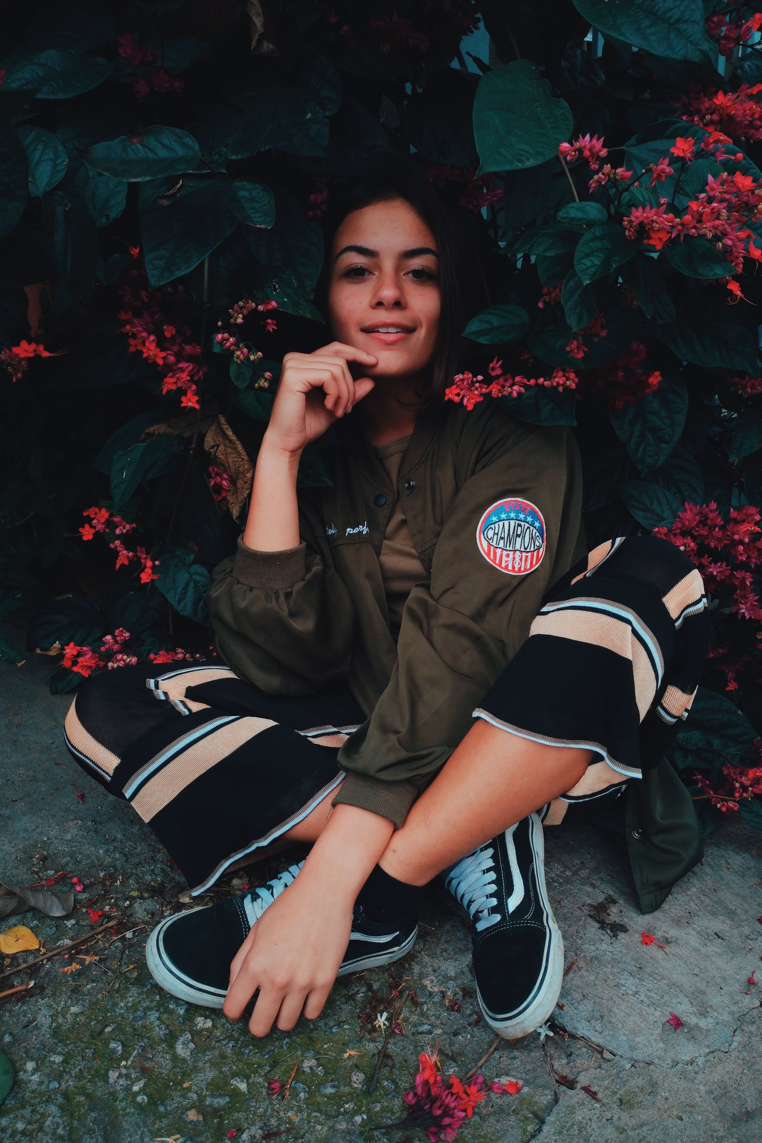 A smiling woman in casual attire sits amidst vibrant foliage, presenting a relaxed and natural vibe.