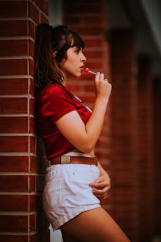 Profile view of a young woman savoring a lollipop while leaning against a brick wall.