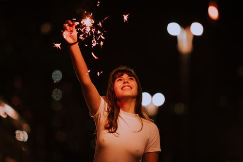 Free A woman joyfully holds a sparkler under the night sky, surrounded by bokeh lights. Stock Photo