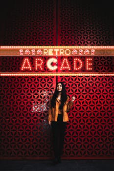 Woman standing under illuminated retro arcade neon sign on patterned red wall.
