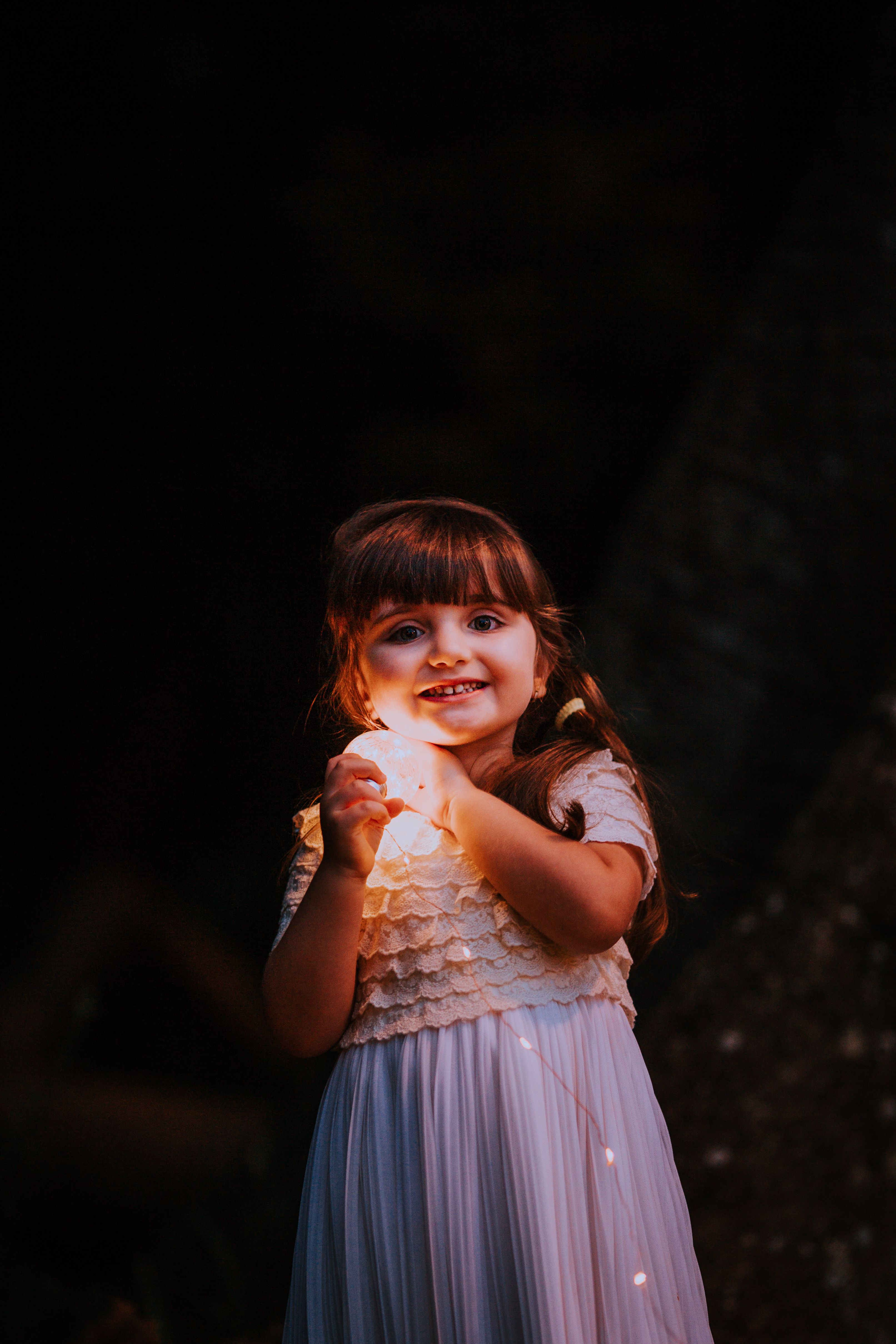 A young girl in a white dress smiling warmly while holding twinkling string lights outdoors.