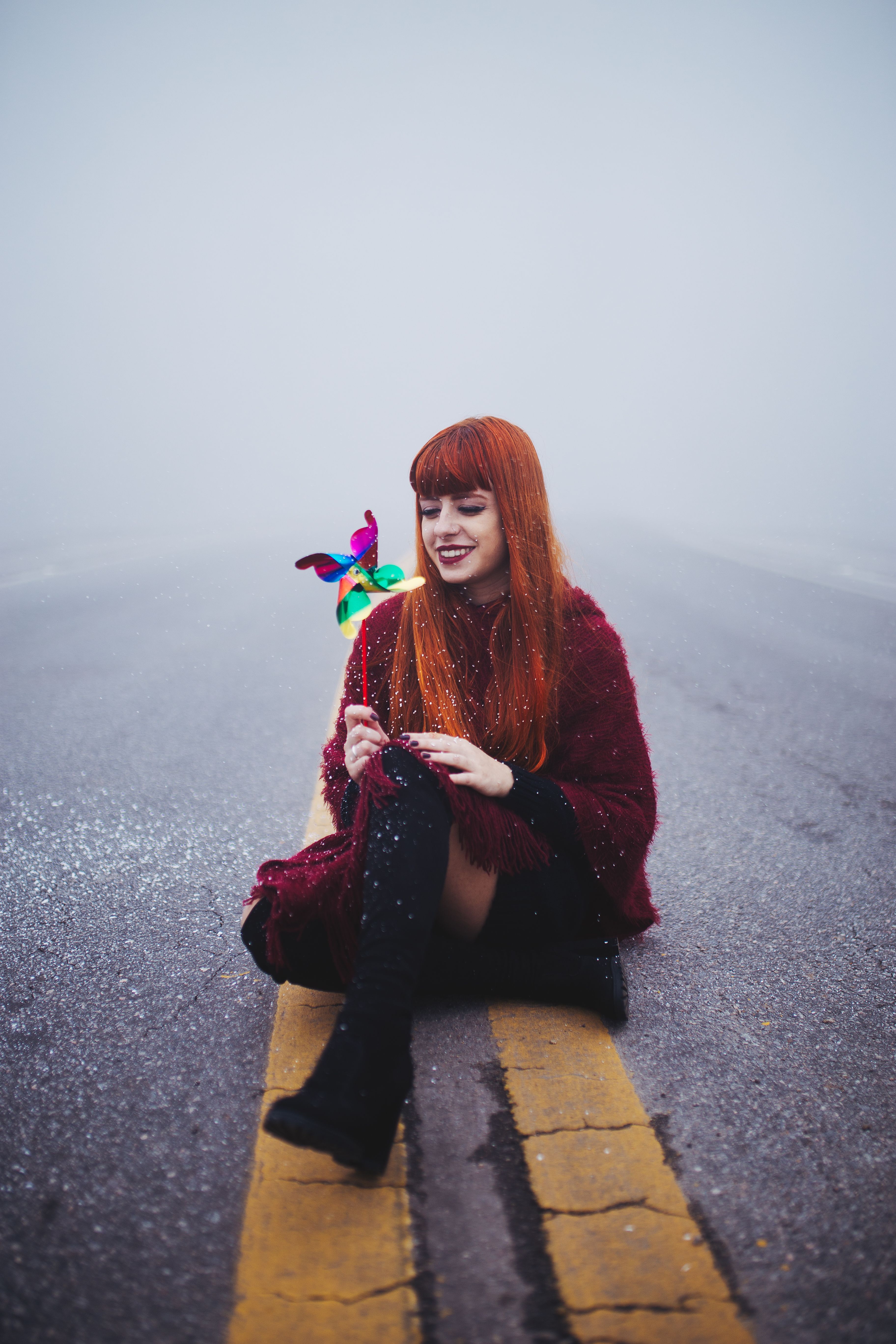 A redhead woman with a pinwheel sits smiling on a snowy road during winter.