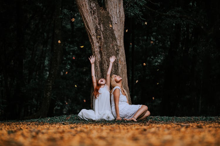 Girl And Woman In Dresses Sitting By Tree