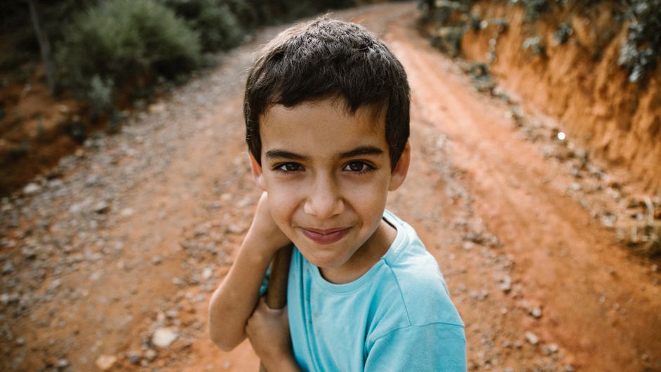Un niño alegre con una camisa azul sonríe cálidamente en un camino de tierra rodeado de naturaleza.