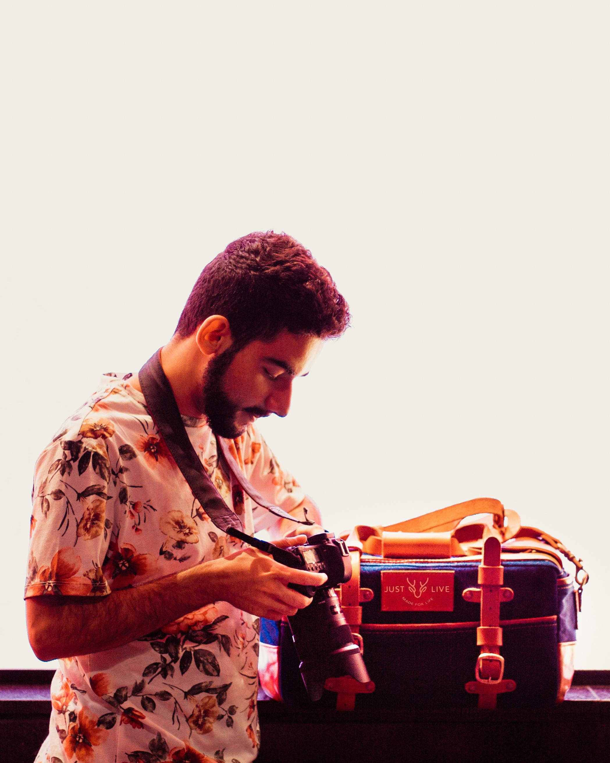 A man in a floral shirt inspects his DSLR camera beside a stylish travel bag indoors.