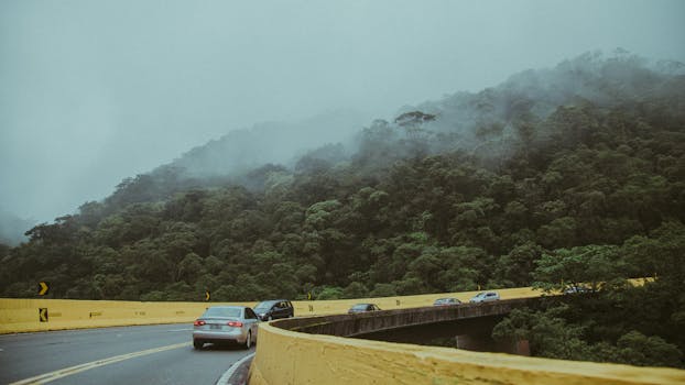 A scenic foggy road winding through lush green mountains, featuring multiple cars, mist, and forest.