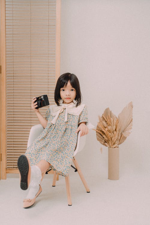 A Girl in Floral Dress Sitting on Chair Holding Camera