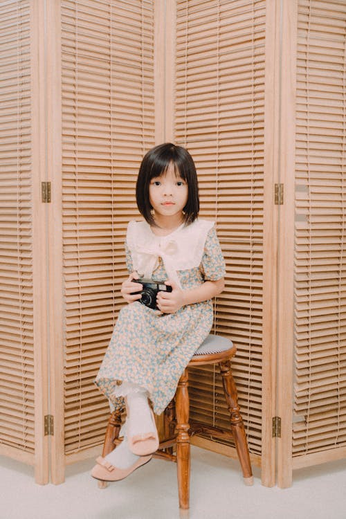 Girl in Floral Dress Sitting on Wooden Chair Holding Camera