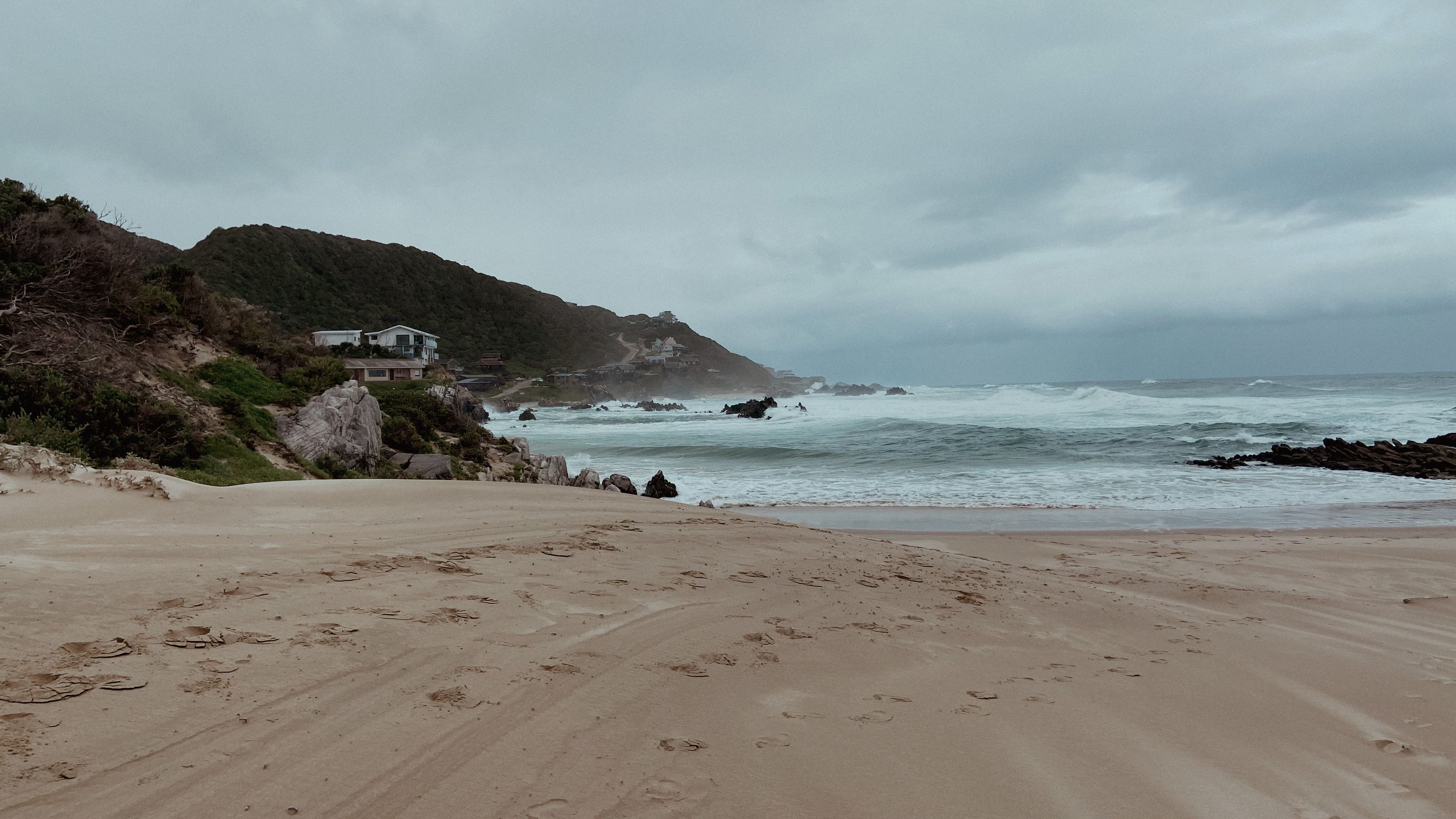 A scenic overcast beach in Kareedouw, South Africa, featuring waves, hills, and coastal houses.