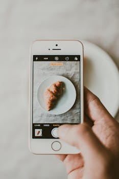 A person photographs a croissant on a plate using a smartphone indoors.