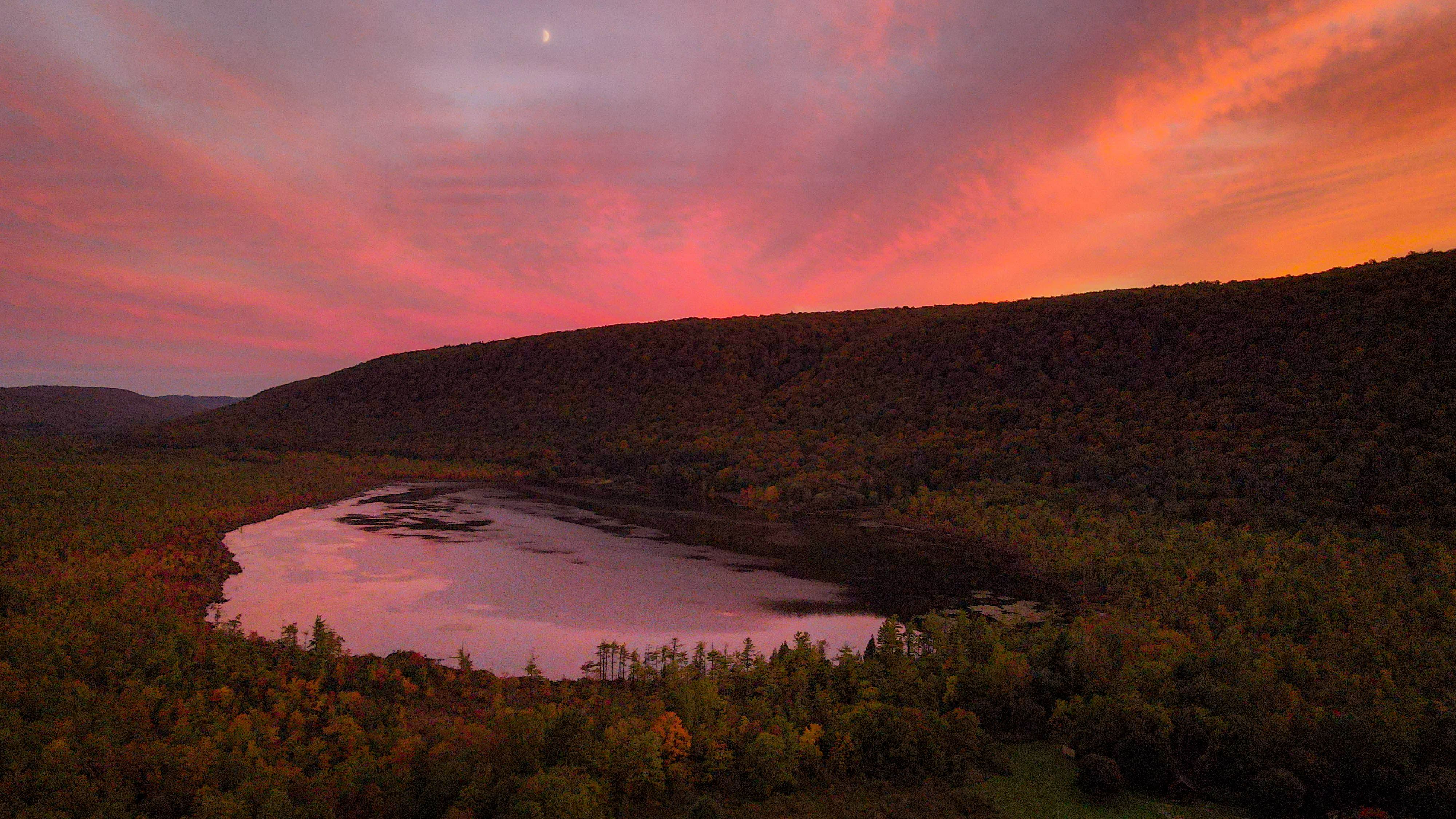 A breathtaking aerial view of an Adirondack lake surrounded by fall foliage under a vibrant sunset sky.
