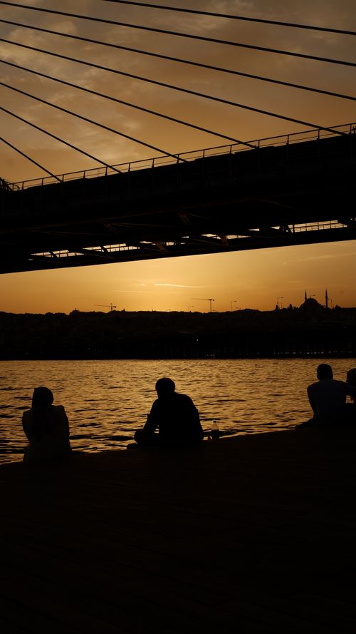 Free People silhouetted against a golden sunset by a bridge over calm waters. Stock Photo