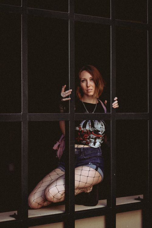 Free A fashionable woman in denim shorts, fishnet stockings, and a printed shirt poses behind a metal frame. Stock Photo