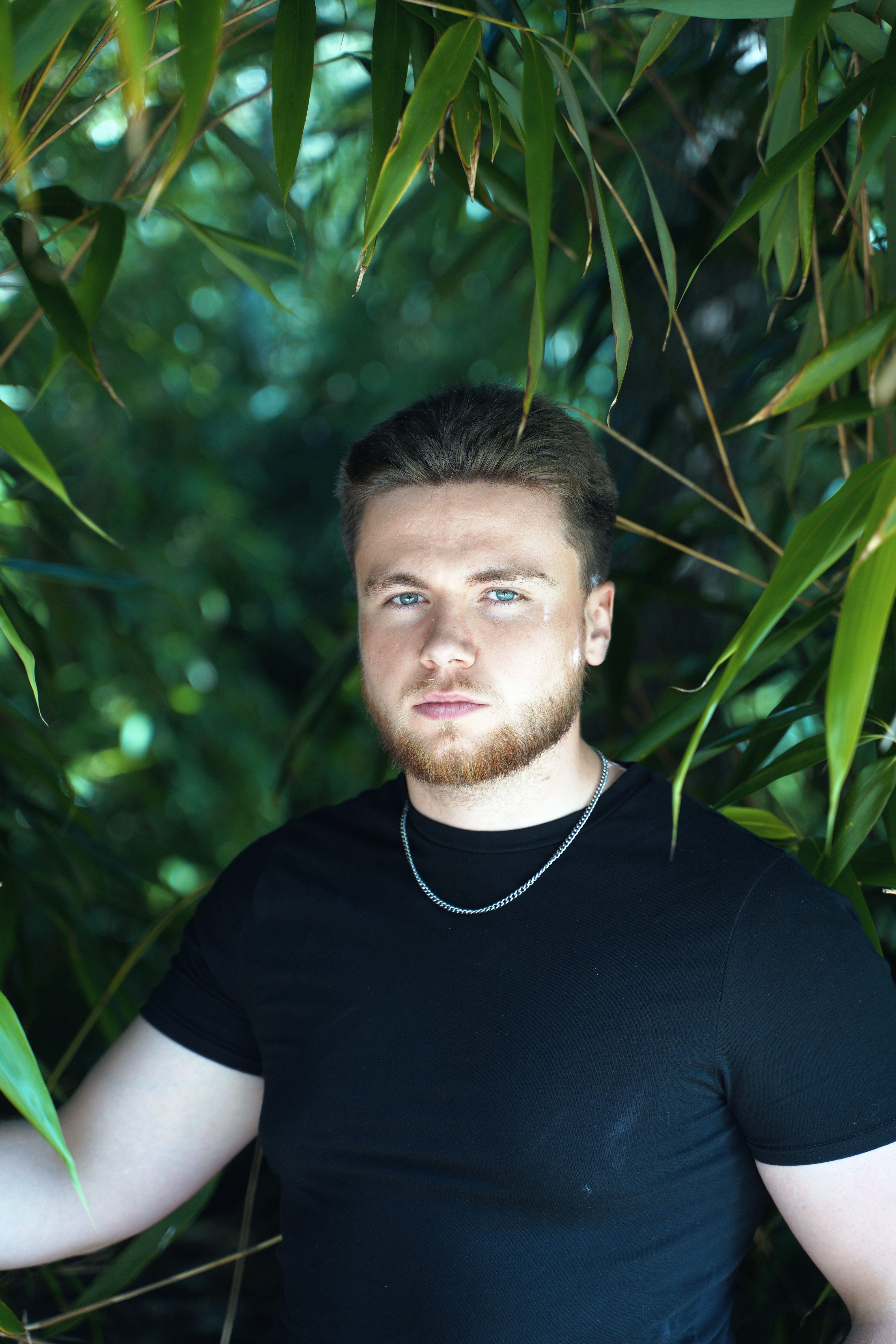 A bearded man in a black shirt poses amidst vibrant green leaves, exuding a serious demeanor.