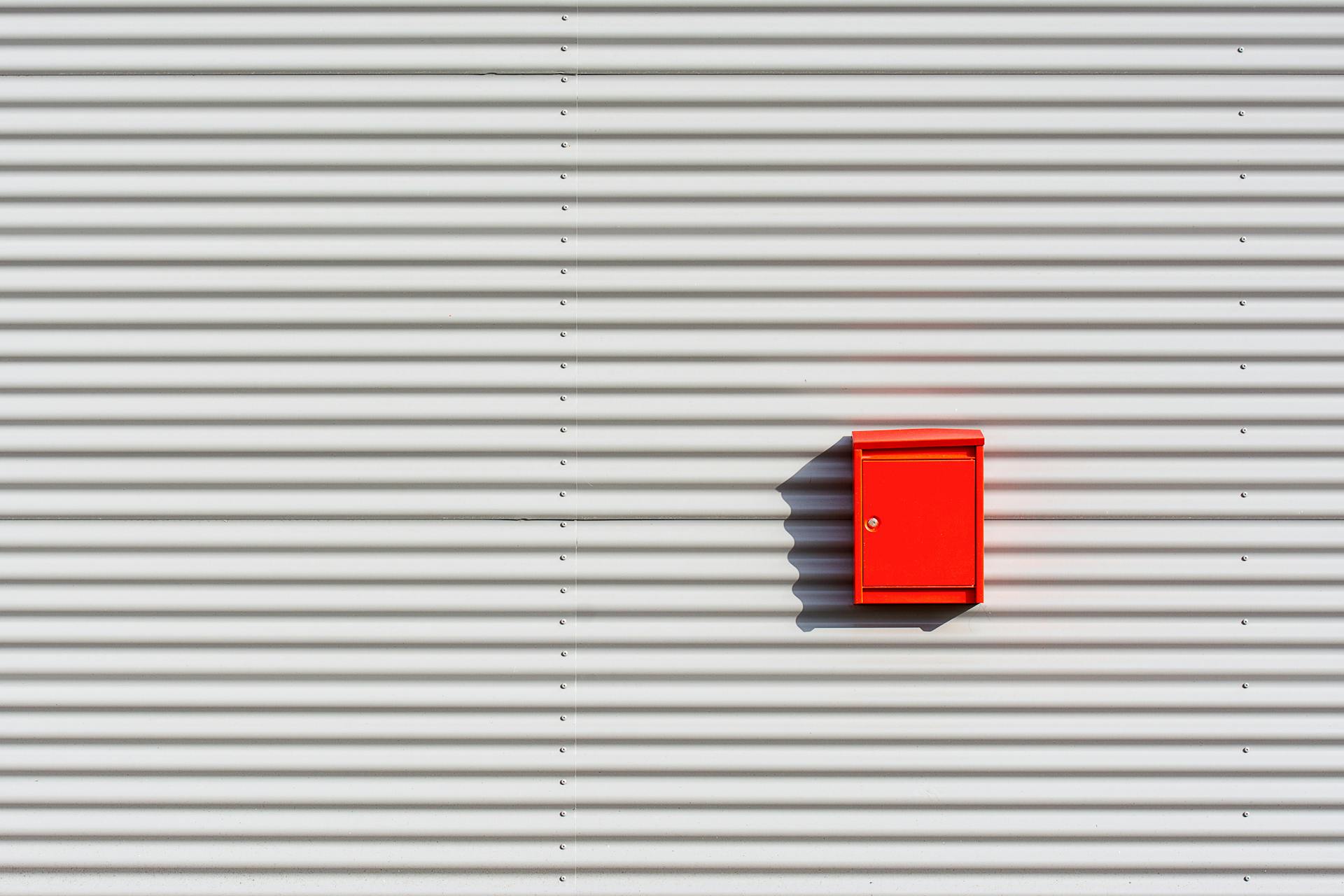 Red mailbox mounted on a minimalist striped white wall casting a dramatic shadow.