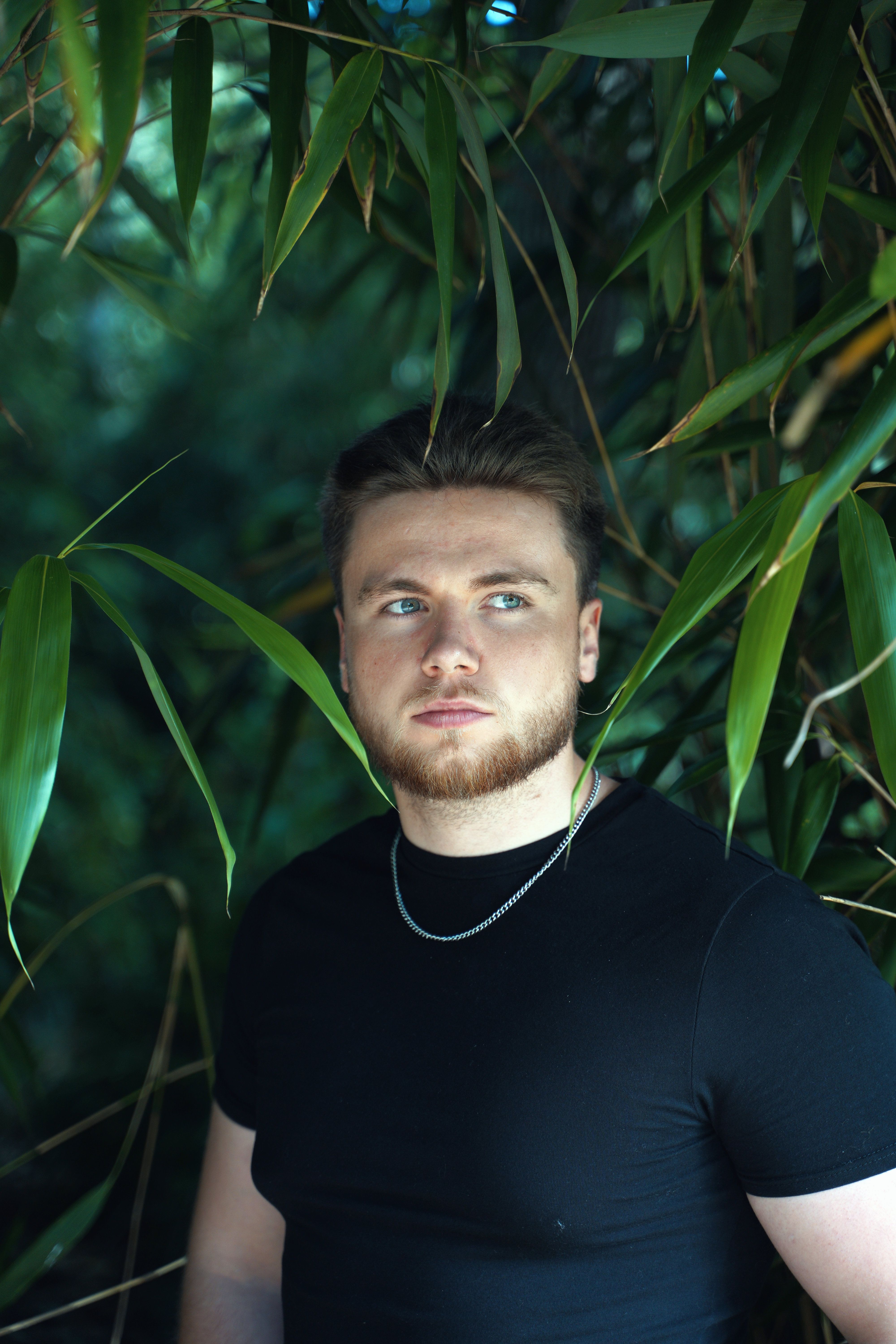 Young man with facial hair wearing a black shirt, surrounded by lush green leaves.