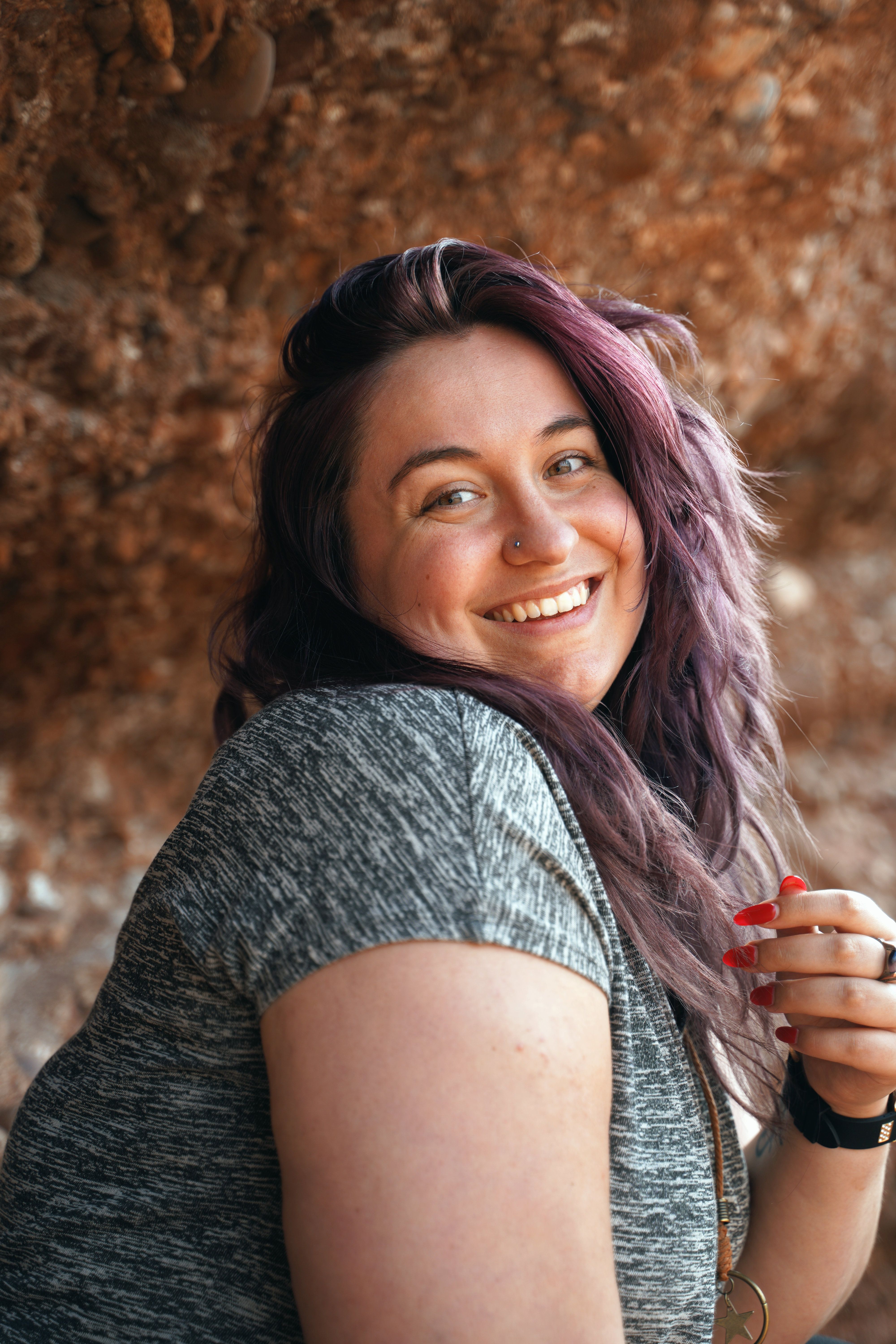 Portrait of a smiling woman with purple hair and a nose piercing in a natural setting.