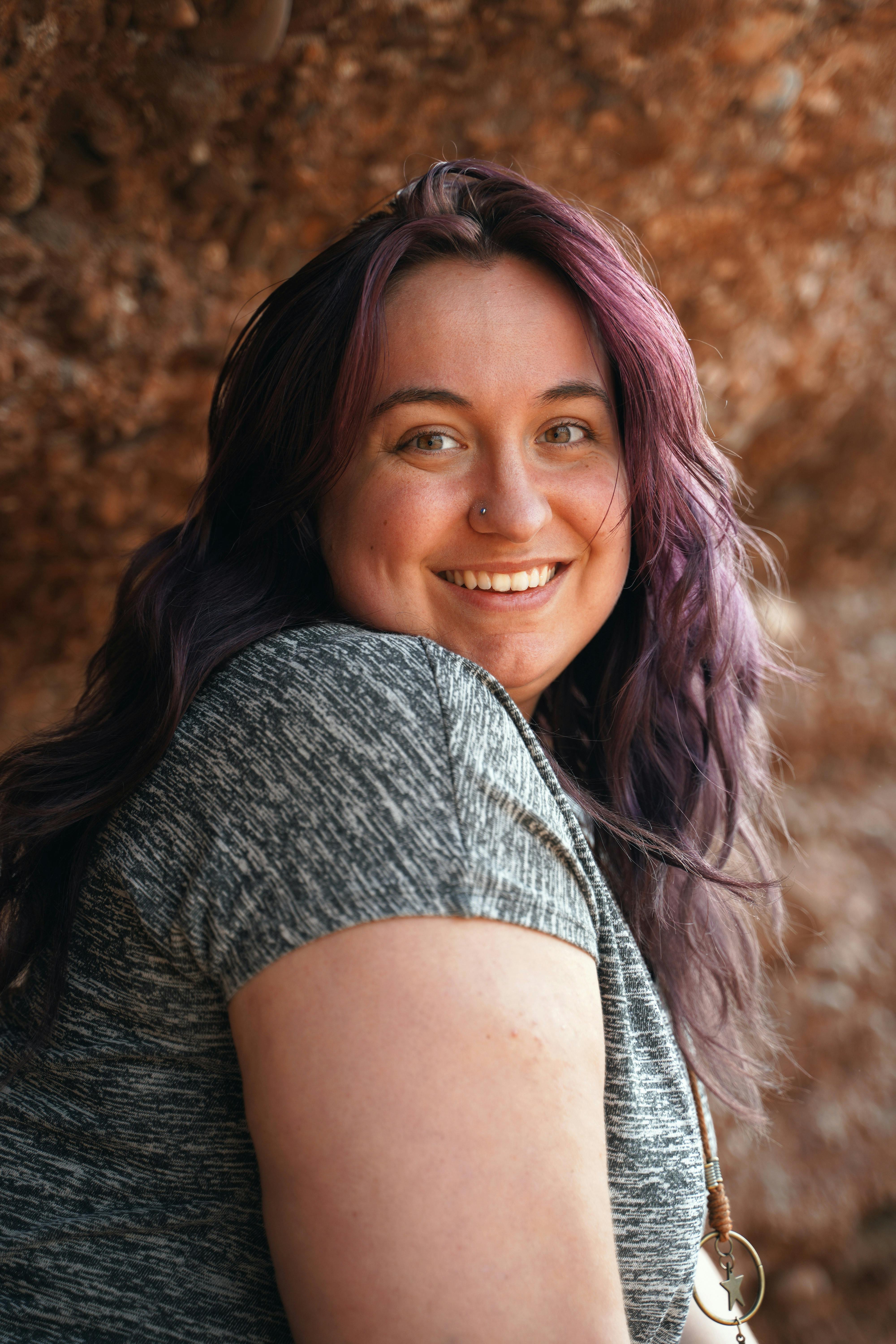 A cheerful woman with purple hair smiling near a rock formation, radiating happiness.
