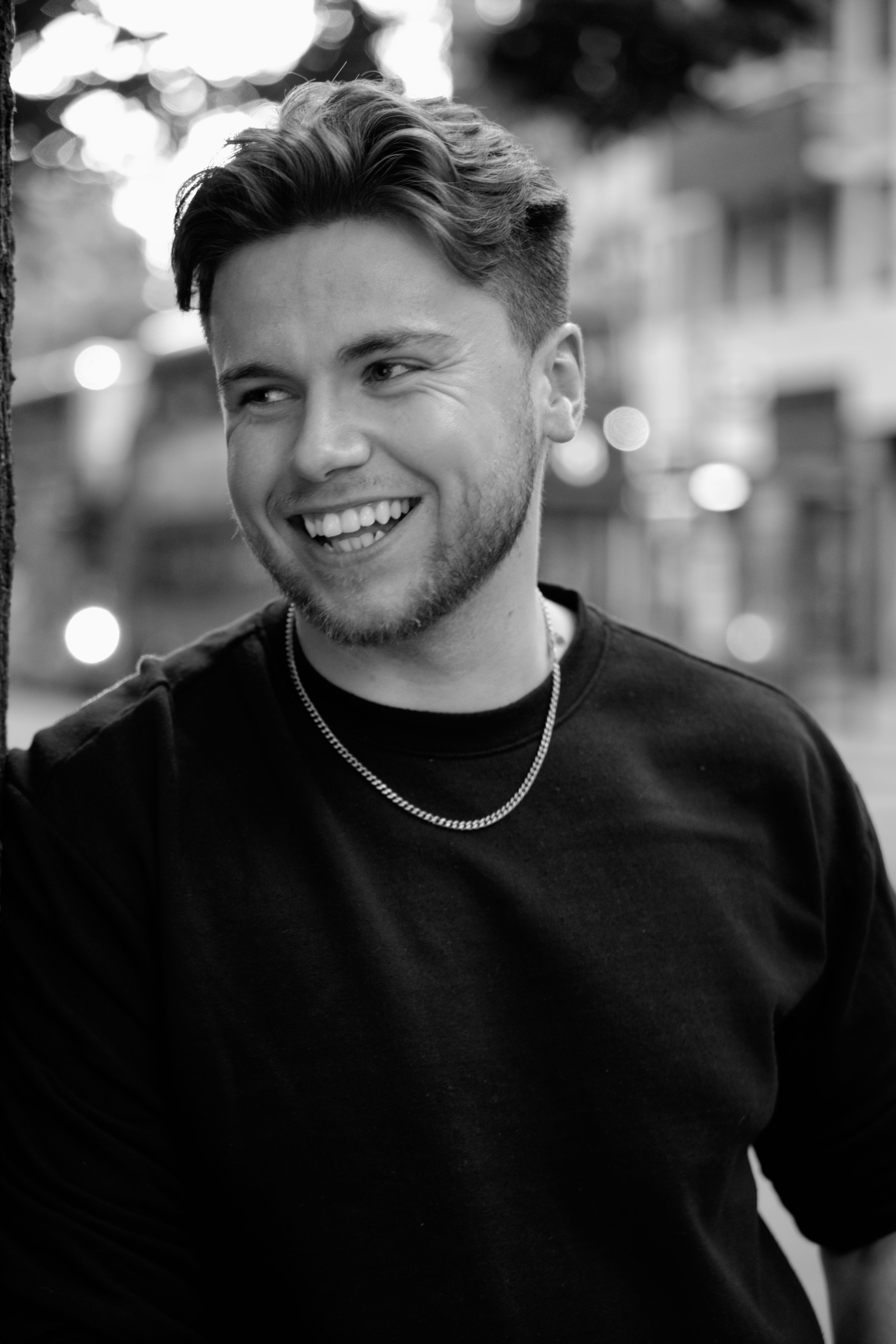 Black and white photo of a smiling man in casual attire with bokeh background outdoors.