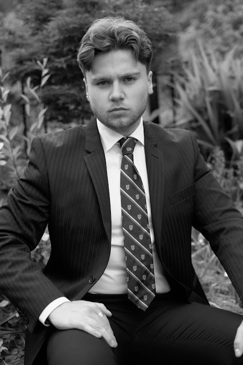 A Grayscale Photo of a Man in Black Suit Sitting while Looking with a Serious Face
