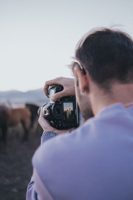 A photographer in a purple sweater captures a scenic view of horses using a DSLR camera.