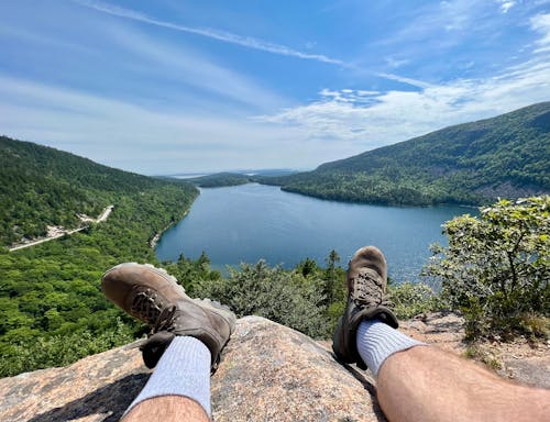 Foto de archivo gratuita de una persona en pantalones cortos negros y zapatos de senderismo marrones sentada en una roca cerca de un lago
