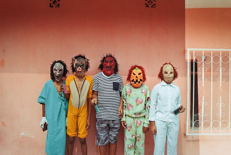 Group Of Children In Masks And Costumes Posing Near House Wall