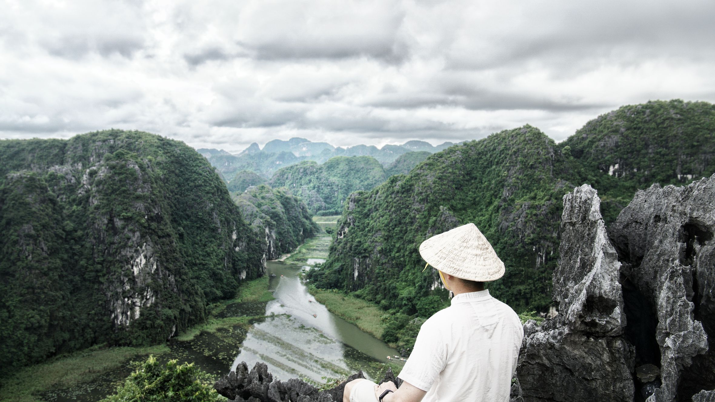 A man with a conical hat sits atop a cliff, overlooking the lush Trang An valley and river in Vietnam.