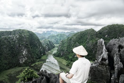 A man with a conical hat sits atop a cliff, overlooking the lush Trang An valley and river in Vietnam.