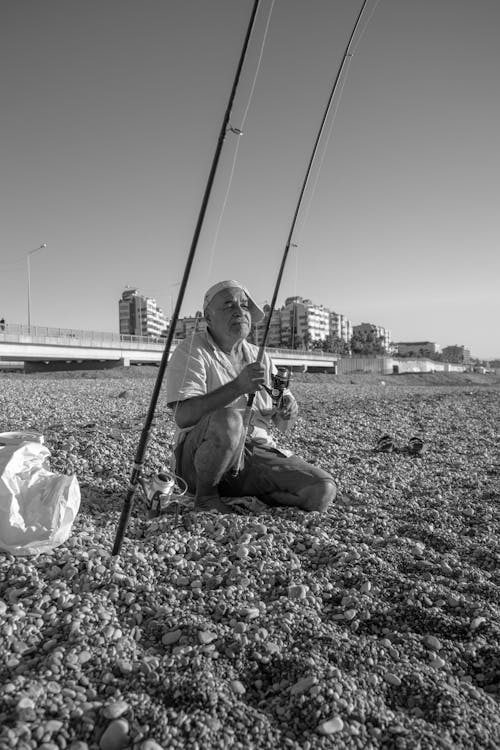 Grayscale Photography of an Elderly Man Sitting on the Shore while Holding a Fishing Rod