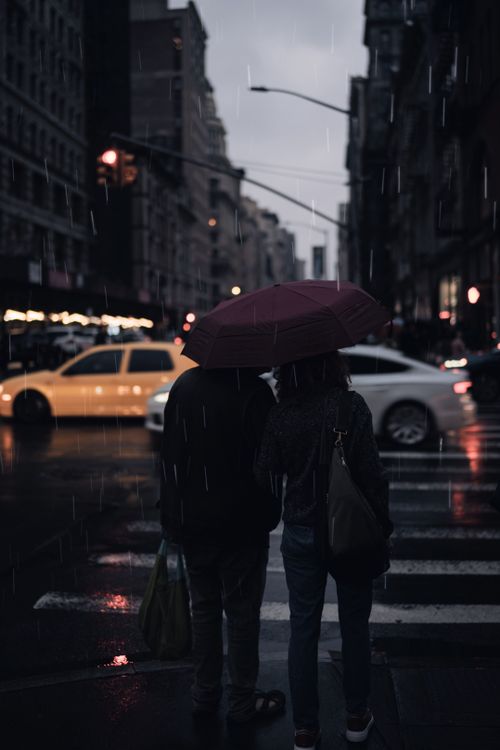 Free A couple stands under an umbrella on a rainy city street, surrounded by cars and buildings. Stock Photo