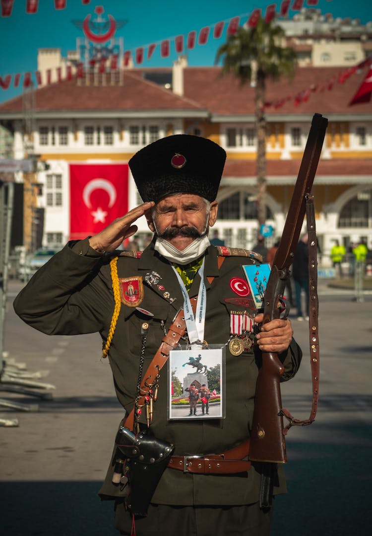 Veteran Wearing A Military Uniform Saluting Holding A Rifle, And Turkish Flag In Background