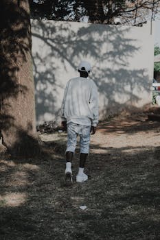 A young man wearing casual attire walks along a shaded outdoor path.