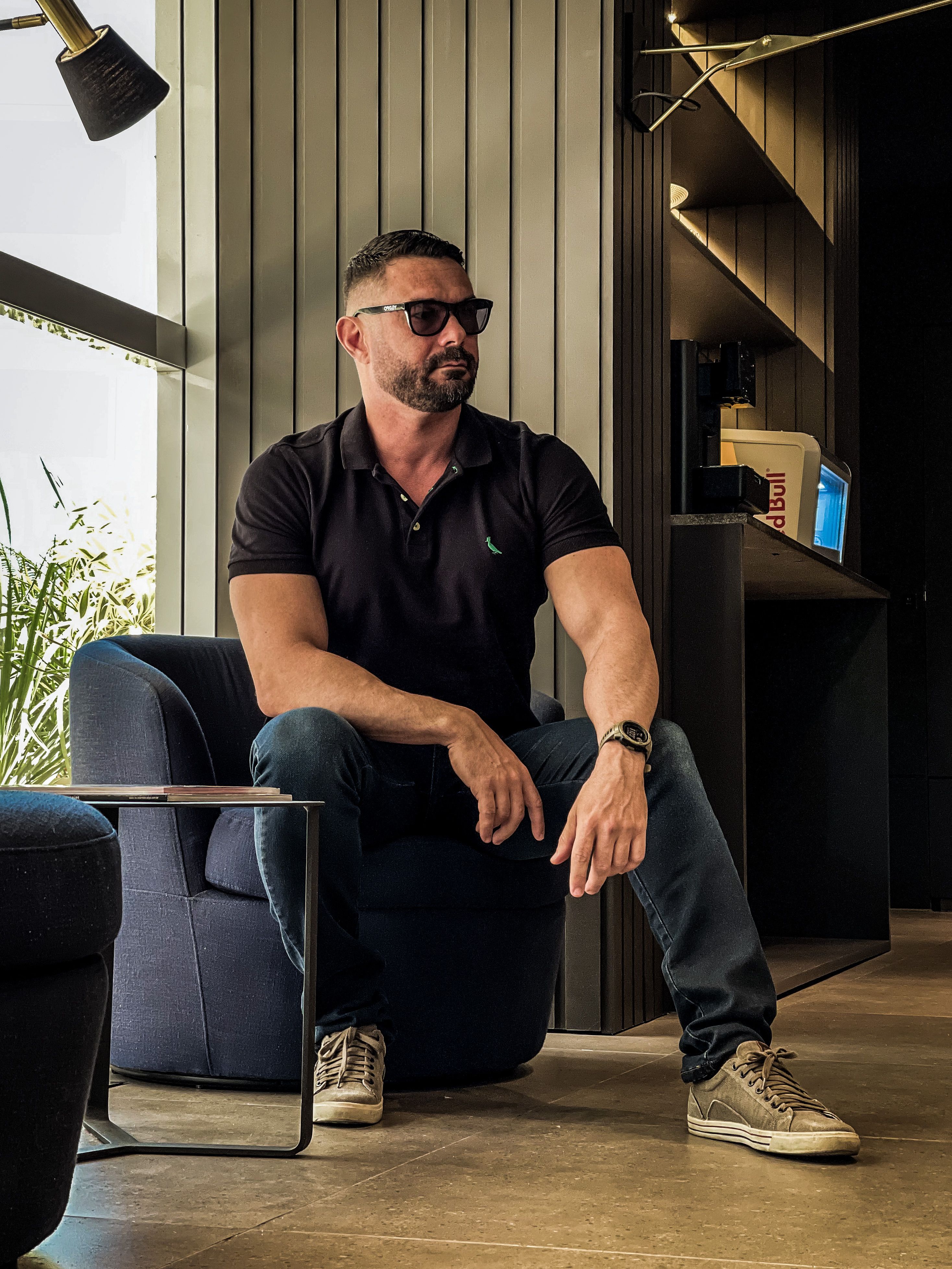Homem barbado em polo preto e jeans, sentado em uma sala moderna usando óculos de sol.
