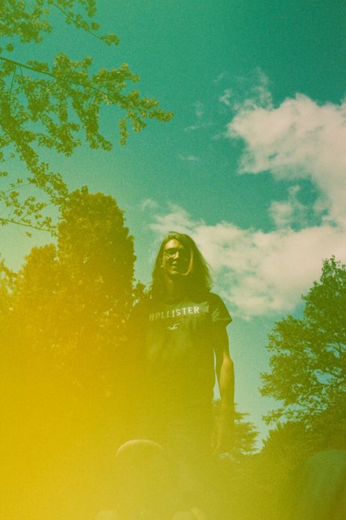 Free A young man with long hair poses with a skateboard under a vibrant summer sky. Stock Photo
