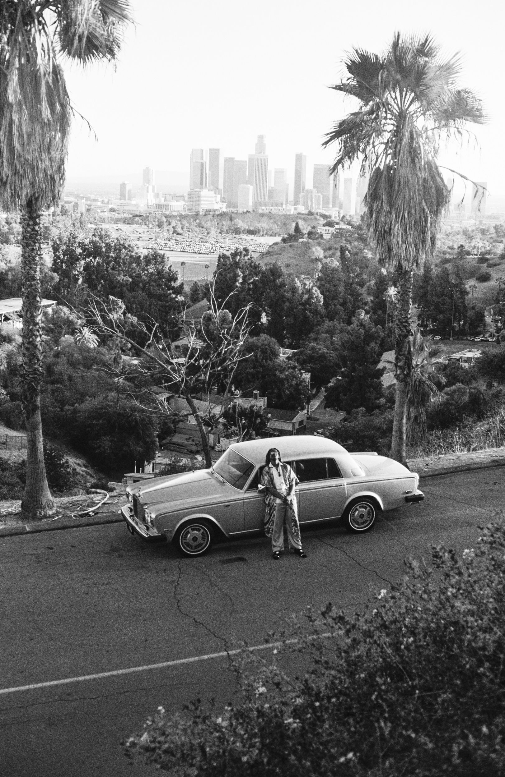 Black and white photo of a vintage car with a man, palm trees, and Los Angeles skyline.