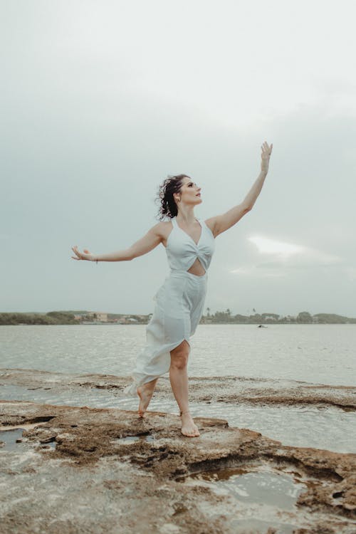 Woman in White Dress Standing on Brown Rock Near Body of Water