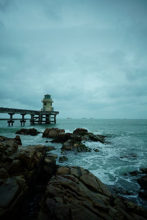 Free A dramatic image of a lighthouse on a rocky shore under a cloudy sky, facing a stormy sea. Stock Photo