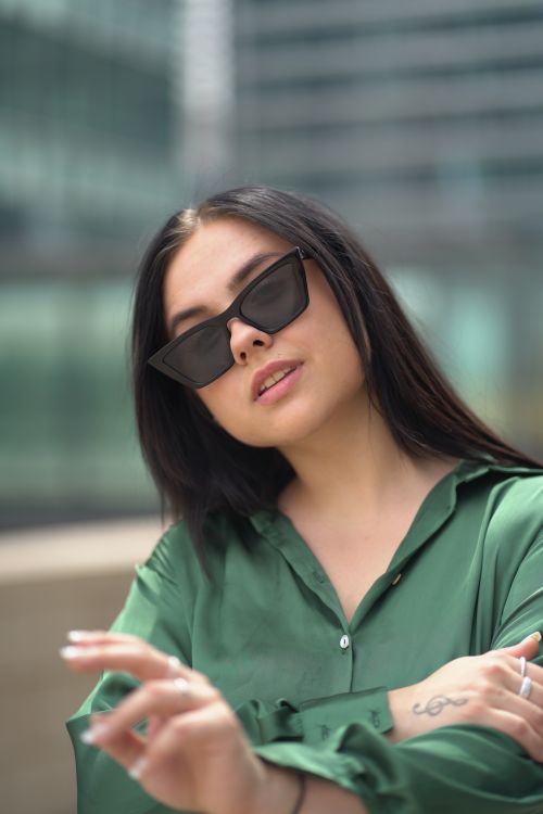 Free Stylish woman in sunglasses wearing a green top poses confidently in a cityscape. Stock Photo