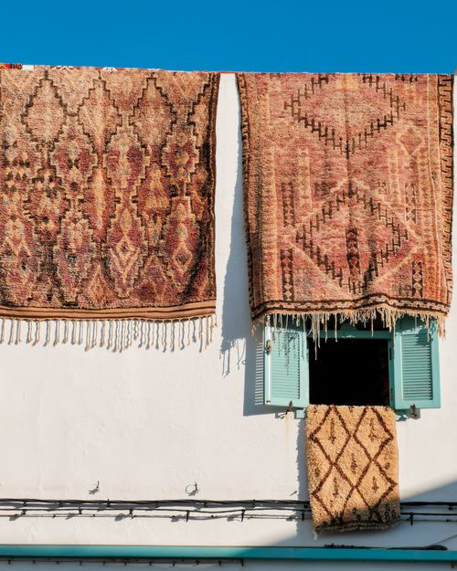 Free Beautiful ethnic handmade rugs hanging on a white wall with blue windows, under clear sky. Stock Photo