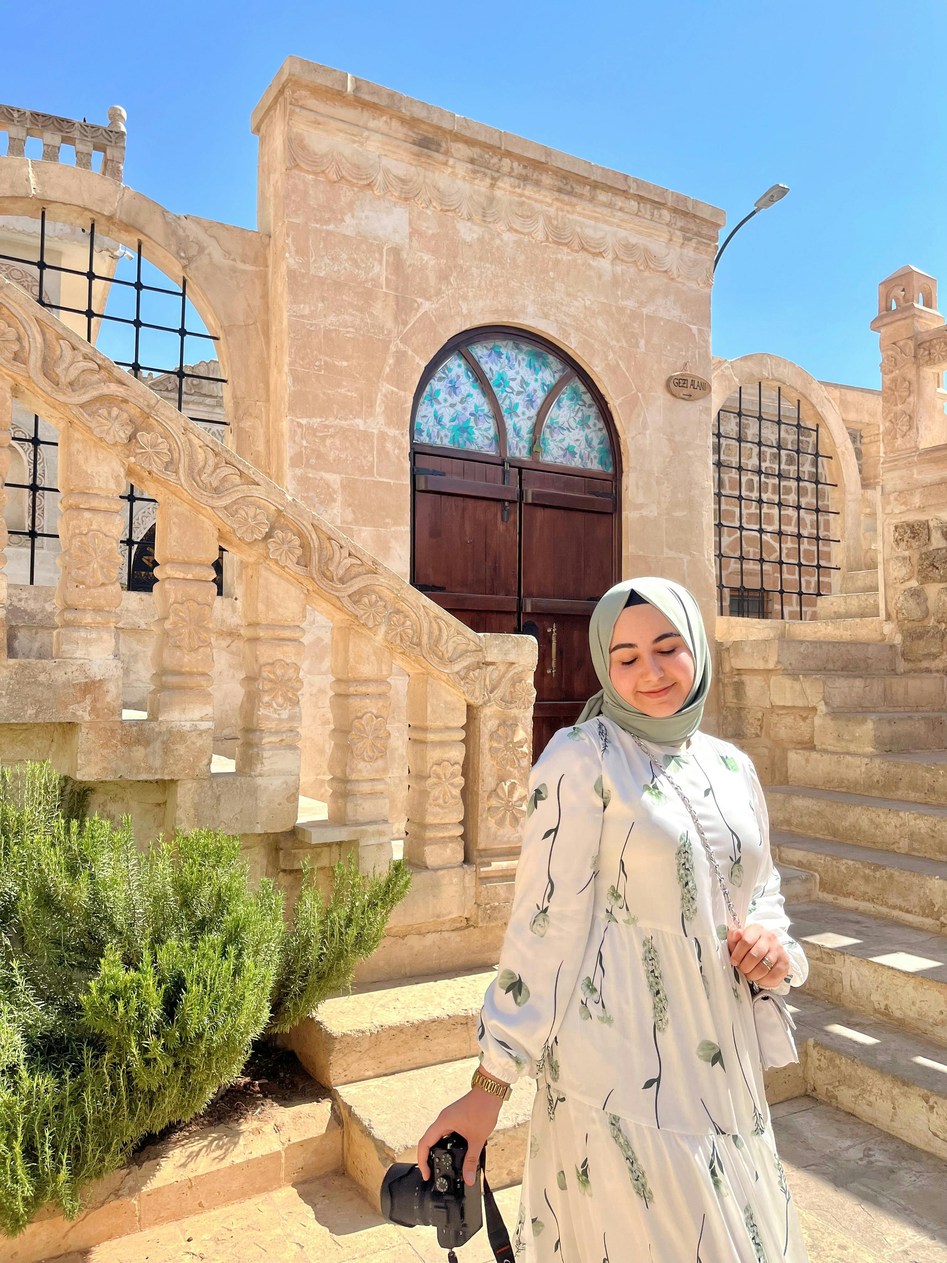 Arabic woman in traditional attire holding a camera outside an ornate stone building.
