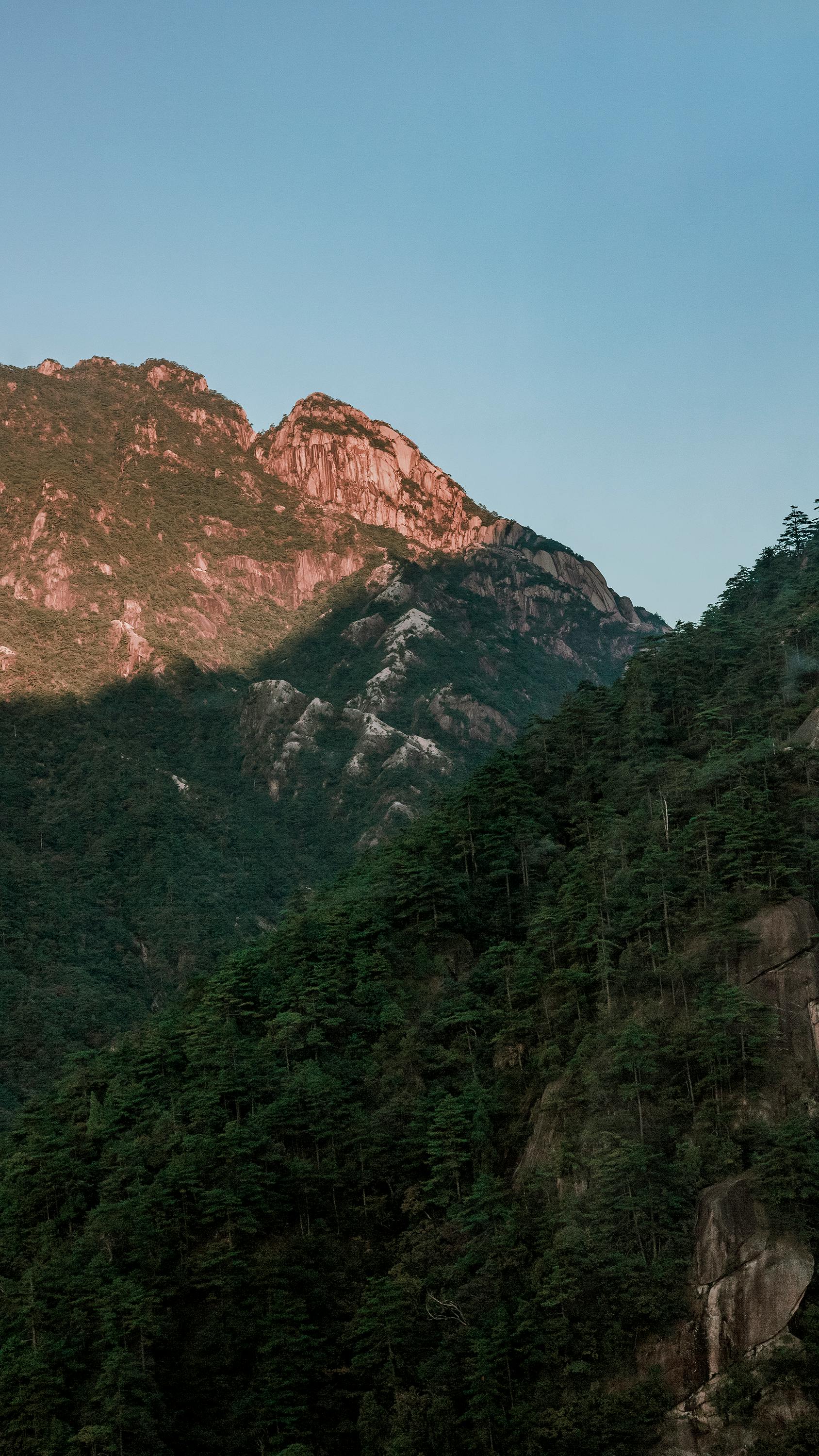 Beautiful rock mountains with tree-covered slopes under a clear blue sky during sunset.