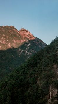Beautiful rock mountains with tree-covered slopes under a clear blue sky during sunset.