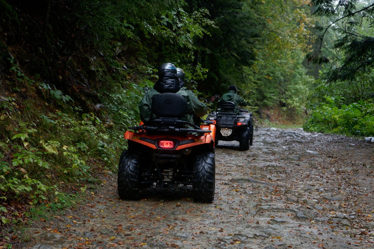 Two ATV riders navigating muddy forest trail wearing proper off-road riding gear and helmets